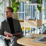 confident-young-business-coach-holds-a-book-and-looks-away-sitting-at-a-table-with-laptop-in-office.jpg